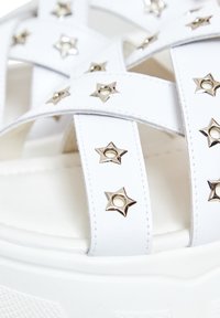 Bílé platformové sandály s křížovými popruhy, ozdobené stříbrnými hvězdicovitými nýty. Hladký povrch a polstrovaná podrážka pro pohodlí.