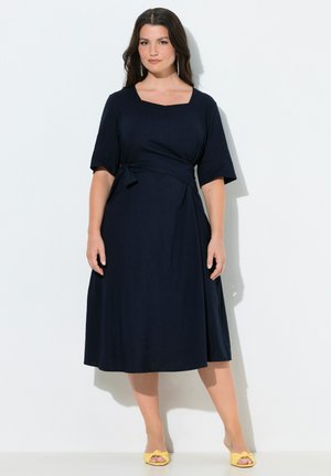 Mujer con un vestido azul marino hasta la rodilla, de media manga y cintura atada, combinando con tacones amarillos abiertos, de pie contra una pared blanca.