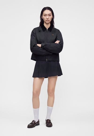 Femme debout, bras croisés, portant une veste zippée noire, une jupe plissée noire, des chaussettes blanches et des mocassins marron foncé sur un fond uni.