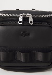 Sac synthétique noir avec texture lisse, doubles fermetures éclair argentées et logo noir en relief. Comprend un accent horizontal en tissu.