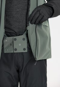 Grøn jakke med sort foring, der har trykknaplukning og lynlås, sammen med sorte bukser. Handsker dækker hånden.