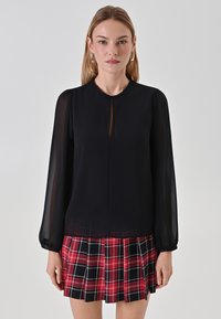Schwarze Bluse mit langen Ärmeln, durchsichtigen Ärmeln und Schlüssellochausschnitt, kombiniert mit einem roten und schwarzen karierten Faltenrock.