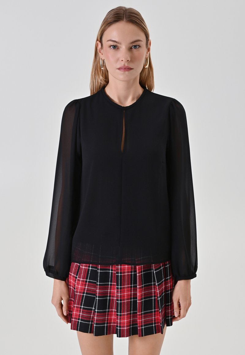 Schwarze Bluse mit langen Ärmeln, durchsichtigen Ärmeln und Schlüssellochausschnitt, kombiniert mit einem roten und schwarzen karierten Faltenrock.
