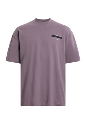 Camiseta de manga corta y cuello redondo en lavanda apagada, hecha de algodón. Presenta una etiqueta de logo negra en el pecho con detalles de texto en blanco.