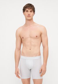 Boxeri din bumbac alb, cu un design ajustat și un elastic cu logo. Modelul este fără tricou, pe un fundal neutru.