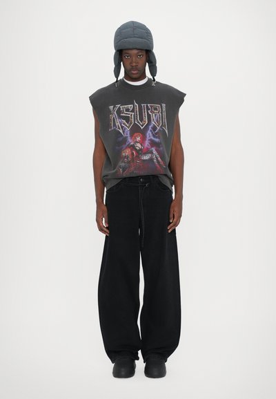 Tricou graphic negru oversized fără mâneci cu logo-ul "KSUBI"; asociat cu pantaloni negri din denim largi și o pălărie gri cu umplutură.