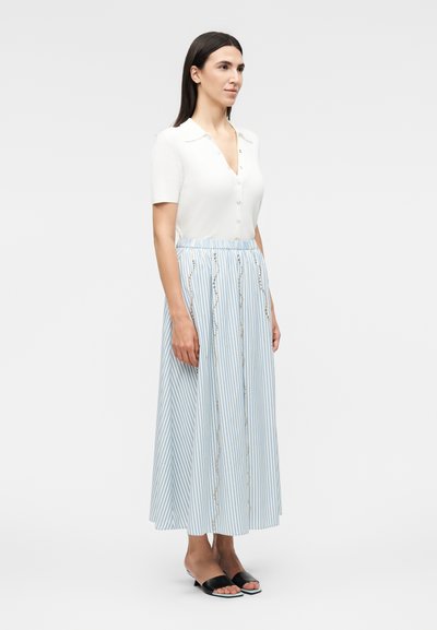 Γυναίκα με μακριά σκούρα μαλλιά που φορά λευκό κοντομάνικο τοπ με κουμπιά, μακριά φούστα μπλε και λευκές ρίγες με σχέδια, και μαύρα παπούτσια με ανοιχτές μύτες.