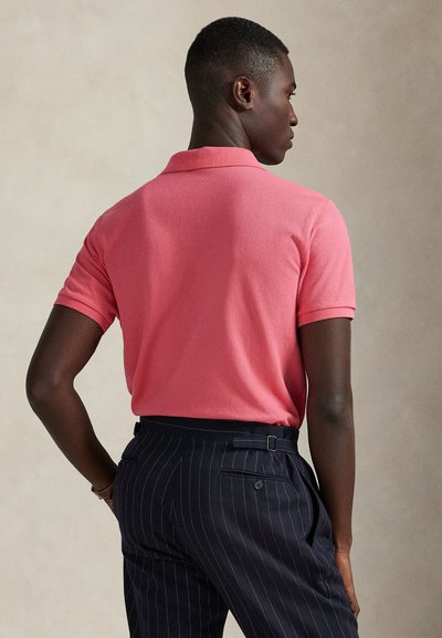 Uomo che indossa una polo rosa corallo a maniche corte e pantaloni blu navy a righe sottili, in piedi con una mano sui fianchi, rivolto di spalle.