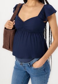 Top peplum de color azul marino con mangas cortas acampanadas. Presenta un detalle fruncido en el busto. Combinado con jeans de color azul claro y un bolso tote marrón.