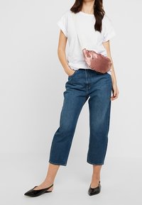 Vrouw in een wit T-shirt, cropped blauwe jeans, zwarte slingbacks en een glanzende roze crossbody tas met witte schouderriem.