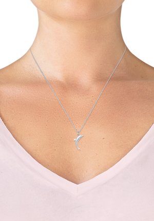 Sølv delfin vedhæng på en tynd kæde, som ligger på en hudfarvet baggrund med en v-hals lys pink top.
