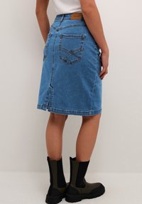 Denim krilo v svetlo modri barvi, do kolen, ravne krojne linije, vidne zadnje žepe in razporek na robu. Usnjena oznaka na pasu.