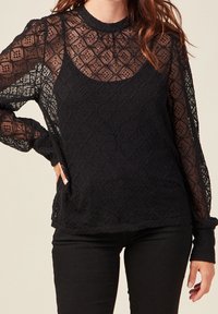 Top lungo nero a maniche lunghe con un design in pizzo testurizzato a motivi a diamante, collo alto e maniche trasparenti, abbinato a uno strato sottostante solido.