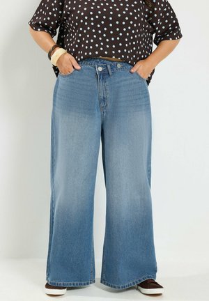 Person iført højttaljede vidde ben blå jeans, brun prikket skjorte og brune sneakers, hænderne i lommerne, stående mod hvid baggrund.