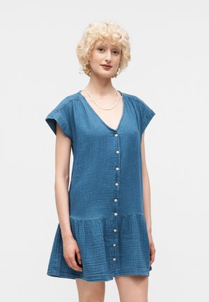 Młoda osoba o kręconych blond włosach, ubrana w niebieską sukienkę z krótkim rękawem, zapinaną na guziki, z obniżoną talią, stojąca na jednolitym tle.