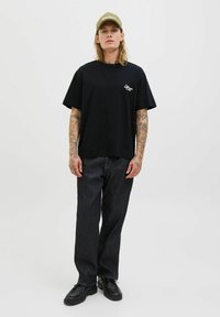 Camiseta de algodón negra con mangas cortas y un logo bordado, combinada con pantalones de mezclilla oscuros y zapatos negros. El modelo tiene el cabello largo y tatuajes.