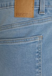 Tecido de denim azul claro com uma textura suave, apresentando um patch de couro castanho rotulado "PIECES" e costura em contraste. Bolso visível.