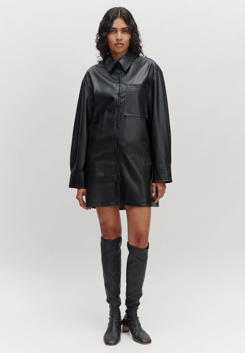 Schwarzes Lederhemdkleid mit langen Ärmeln, Knopfleiste, Brusttasche und glatter Textur. Kombiniert mit hohen schwarzen Stiefeln.