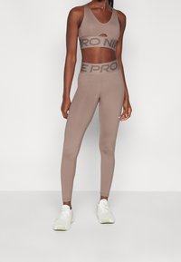 Hnědý sportovní podprsenka a vysoké legíny v setu s výřezy, hladkou texturou a kontrastními logovými pásky. Bílé tenisky.