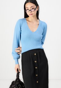 Suéter de malha canelada azul claro com decote em V, combinado com uma saia preta que apresenta seis botões redondos na frente. Mala preta com padrão floral.