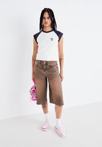 Hvid T-shirt med sorte kortærmer og grøn grafik, brune oversized denimshorts, lyserøde slidesandaler, holder lyserøde sneakers.