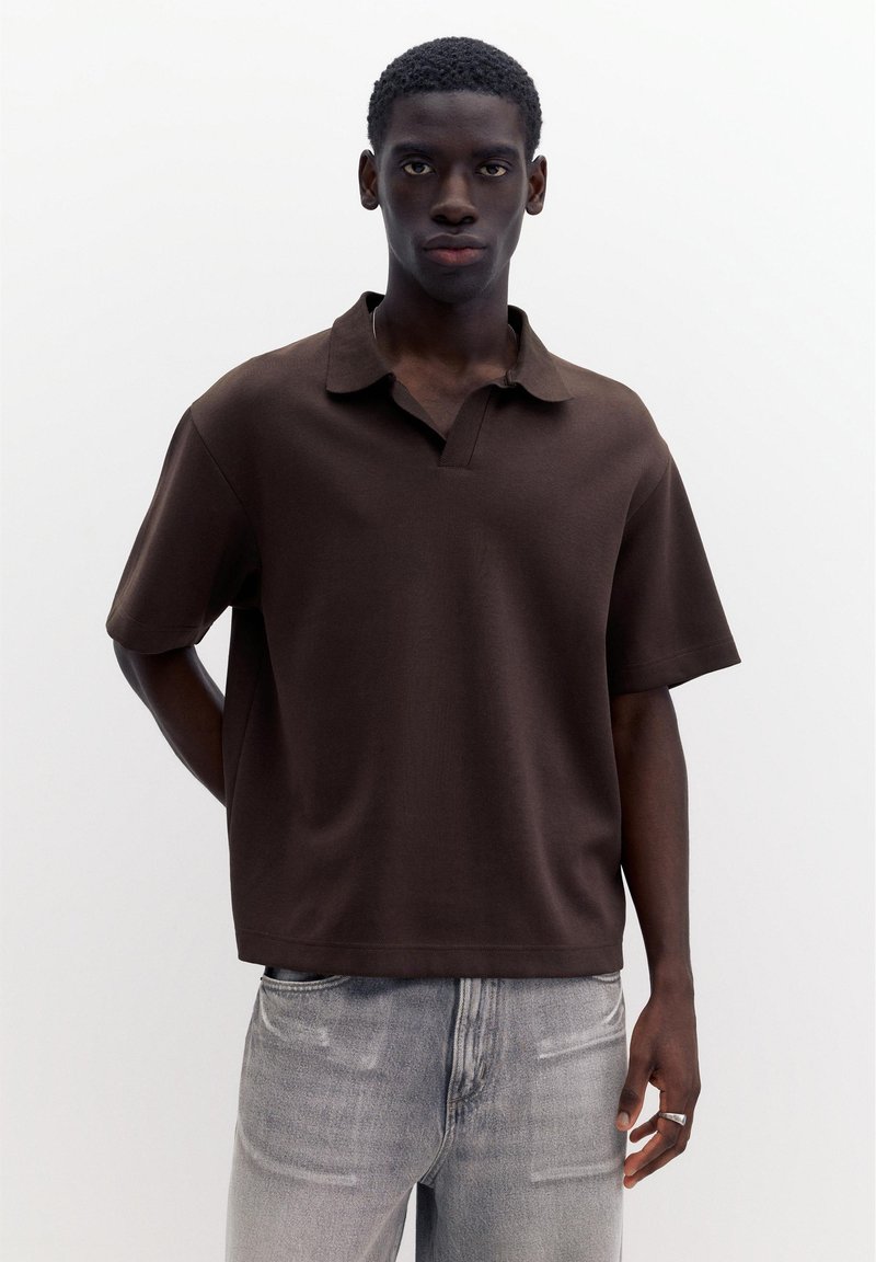 Jeune homme aux cheveux courts et bouclés, portant un polo marron foncé à manches courtes et un jean gris clair, debout devant un fond blanc uni.