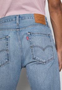 Calças de ganga de denim azul claro com um lavado médio. As características incluem bolsos traseiros, um emblema em pele na cintura e costura em contraste.