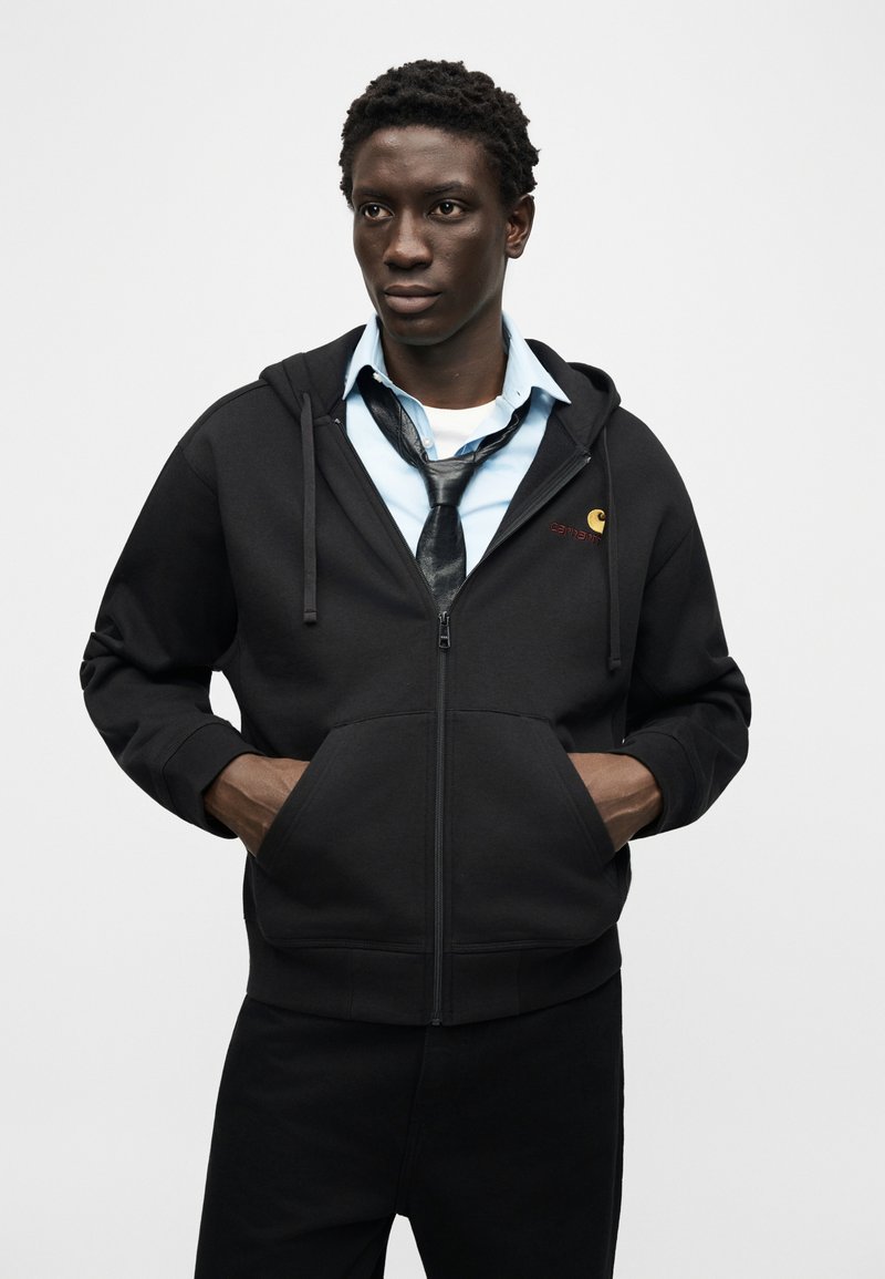 Mann trägt schwarzen Kapuzenpullover mit Reißverschluss über hellblauem Hemd und locker geknüpfter schwarzer Krawatte, steht mit den Händen in den Kapuzenbeuteln vor einfarbigem Hintergrund.