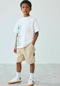 Hvid bomulds t-shirt med blå og grøn grafik "BAKER" på siden, parret med beige cargo shorts og hvide sneakers med gummisåler.