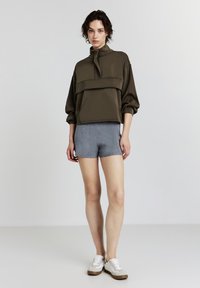 Donna in piedi che indossa un maglione oversize verde oliva, pantaloni shorts di maglia azzurri chiari e sneakers bianche su uno sfondo grigio chiaro uniforme.
