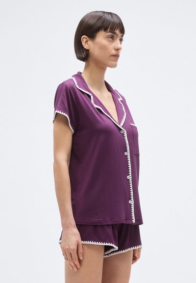 Lila pizsama szett rövid ujjú, fehér csíkozású felsővel és hozzáillő rövidnadrággal, mindkettő puha anyagból készült. Elöl gombos záródással és zsebbel.