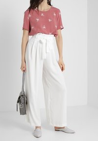 Lyserød top med blomstermotiv, højtaljede hvide bukser med bindebælte, grå håndtaske og lysegrå slips-on sko.