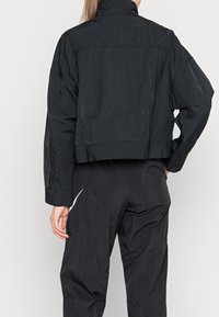 Svart kort jacka i lätt tyg med hög krage och lös passform. Har en jämn textur och subtil glans.