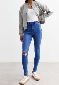 Chaqueta bomber gris corta sobre una camiseta sin mangas blanca, combinada con unos vaqueros ajustados de cintura alta azul con desgastes en las rodillas, y zapatillas blancas con detalles en marrón.