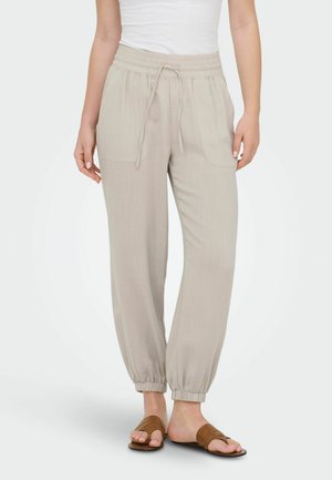 Vrouw draagt beige joggingbroek met trekkoord en elastische boorden en bruine open-teen slippers, staand tegen een effen achtergrond.