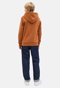 Rust-orange hooded sweatshirt with a ribbed hem, paired with dark blue jeans and white sneakers. Fabric appears soft and casual.