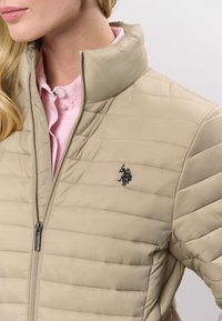 Bēšs polsterēts džekets ar augstu apkakli un horizontālu šūtu dizainu. Uz krūtīm melns izšūts logotips un sudraba rāvējslēdzēja detaļa.