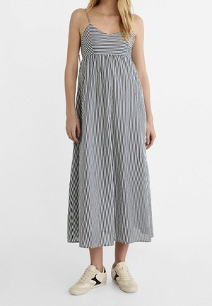 Femme portant une robe midi à rayures verticales noires et blanches avec des bretelles fines et des baskets beiges avec un détail étoile noire.