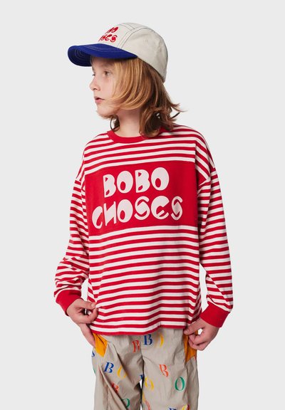 Langärmliges rot-weiß gestreiftes Hemd mit großem weißen "BOBO CHOSES"-Logo; getragen mit einer beigefarbenen und blauen Mütze; hellfarbige gemusterte Hose.