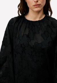 Blouse noire avec un motif floral texturé, fabriquée en tissu léger. Elle comporte un col rond et des détails froncés aux épaules.