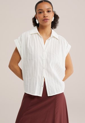 Kobieta o kręconych włosach nosi białą, krótką koszulę z pionowymi paskami i bordową spódnicę, stoi z rękami za plecami.