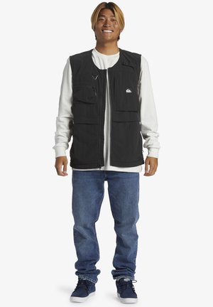 Black, sleeveless vest with multiple pockets and zip closure, worn over a white long-sleeve shirt, paired with blue jeans and dark sneakers.