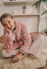 Roosa pehme hommikumantel soonilise tekstuuriga, kantud valge ja roosa triibulise pidžaama komplekti peal. Istub pehmel vaibal, millel paistab hele puidust põrand.