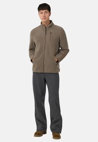 Jonge man in een taupe fleecejack met rits, grijze wijde broek en bruine suède schoenen, staand met handen in de zakken.