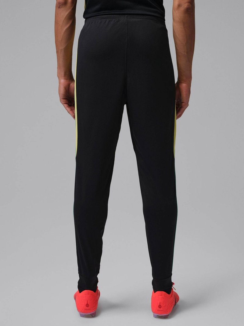 Man die van achteren staat, gekleed in zwarte sportbroek met gele zijstrepen en felrode sportschoenen tegen een grijze achtergrond.