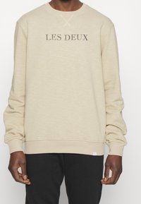 Hombre vestido con sudadera de manga larga beige con el texto "LES DEUX" en el frontal y pantalones negros, de pie contra un fondo blanco.