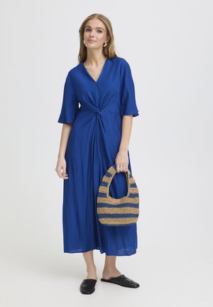 Žena v modrých midi šatách drží pruhovaný tkaný kabelku, má na nohách čierne slip-on topánky, stojí na jednoduchom svetlom pozadí.