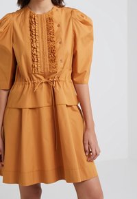 Sukienka peplum w musztardowej bawełnie z bufkami. Posiada pionowe wykończenia w falbany oraz związany w talii, co zapewnia dopasowany wygląd.