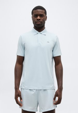 Muž stojaci čelom vpred, oblečený v svetlomodrej polokošeli s krátkym rukávom a zodpovedajúcich šortkách na čistom bielom pozadí.