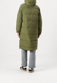 Long olive green puffer coat with vertical quilted panels, featuring a hood and elastic cuffs, worn over light blue jeans and black sneakers.